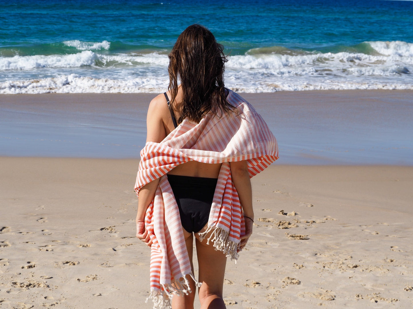 Woman in a coral striped Portsea Turkish cotton beach towel walking on the beach, enjoying the ocean view.