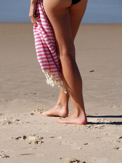 Woman holding Portsea Turkish Cotton Beach Towel in rose stripes on sandy beach, perfect for style and quick drying.