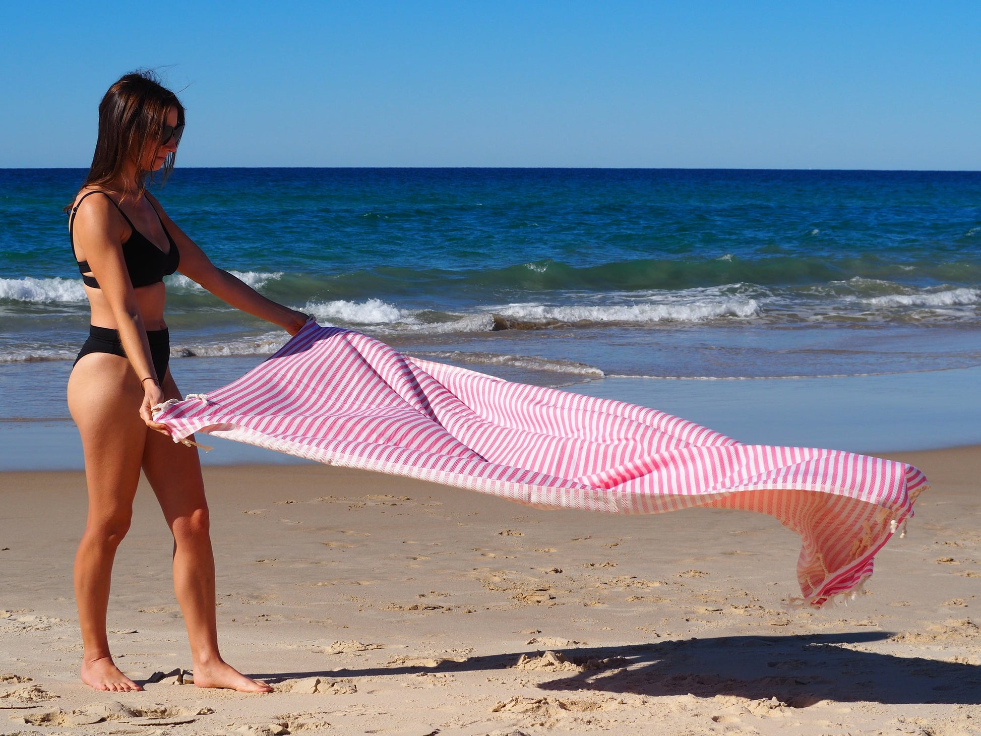 Woman in a bikini holding a Portsea Turkish cotton beach towel on the shore, showcasing its quick-dry feature and style.