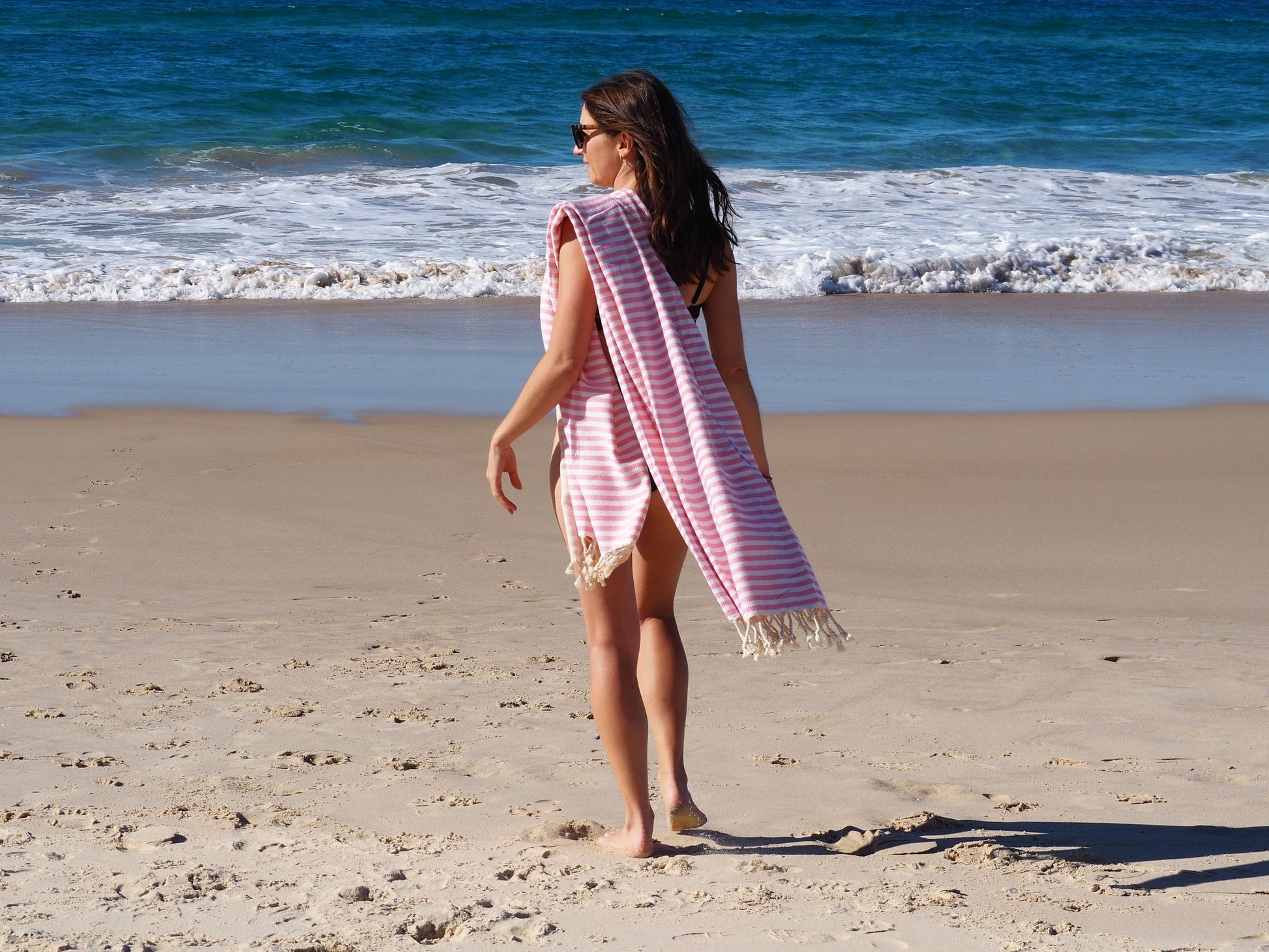 Woman walking on the beach with a blush striped Turkish cotton beach towel, enjoying a sunny day by the water.