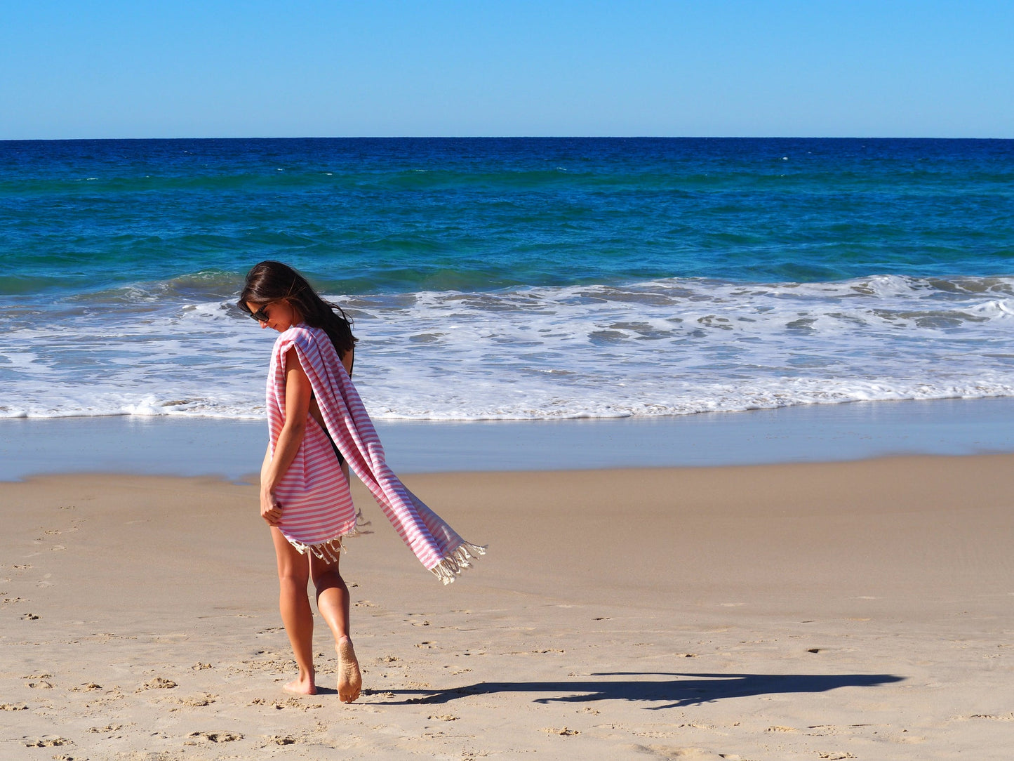 Woman walking on the beach with a blush Turkish cotton towel, enjoying a sunny day by the ocean.