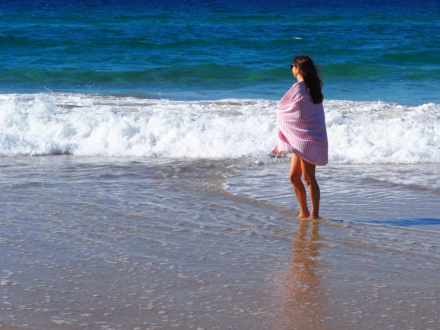 Woman in blush Turkish cotton beach towel standing by the ocean, enjoying a sunny day at the beach.