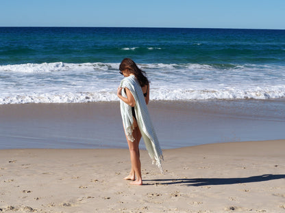 Young woman holding a seafoam Turkish cotton beach towel on the shore, enjoying a sunny beach day.