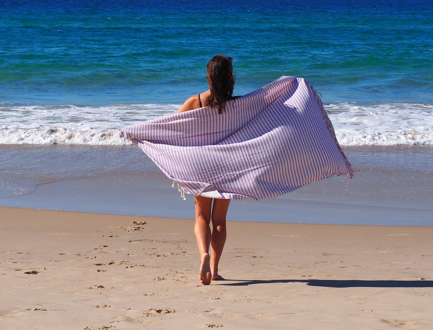 Woman walking on the beach with a lilac striped Turkish cotton beach towel, enjoying a sunny day by the ocean.