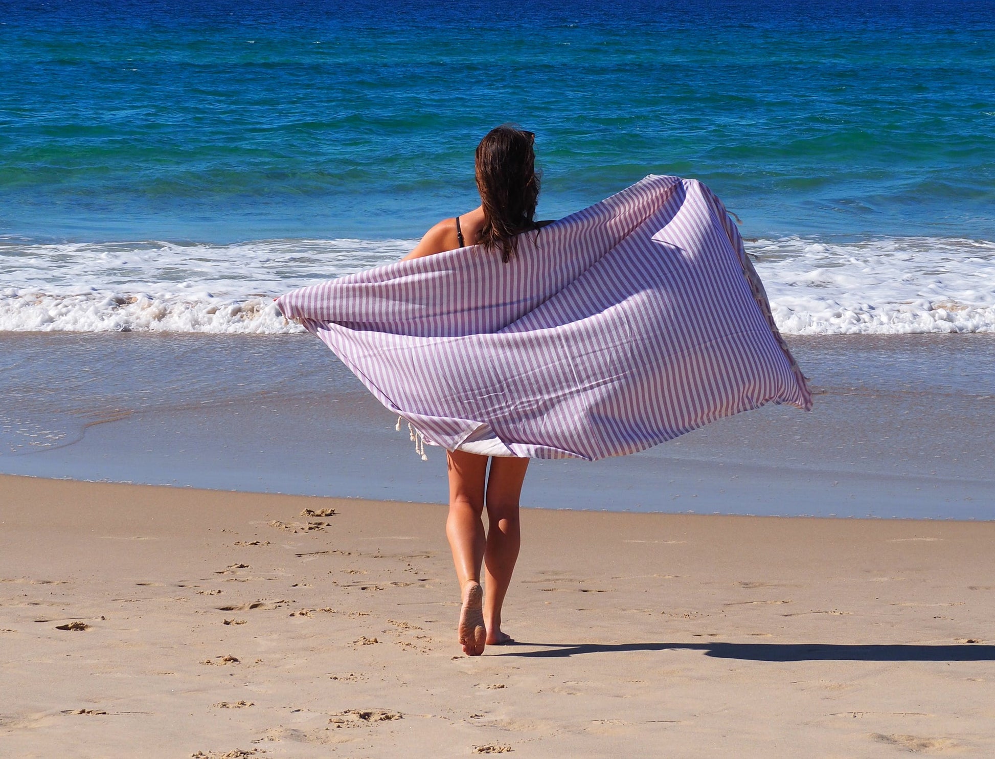 Woman walking on the beach with a lilac striped Turkish cotton beach towel, enjoying a sunny day by the ocean.