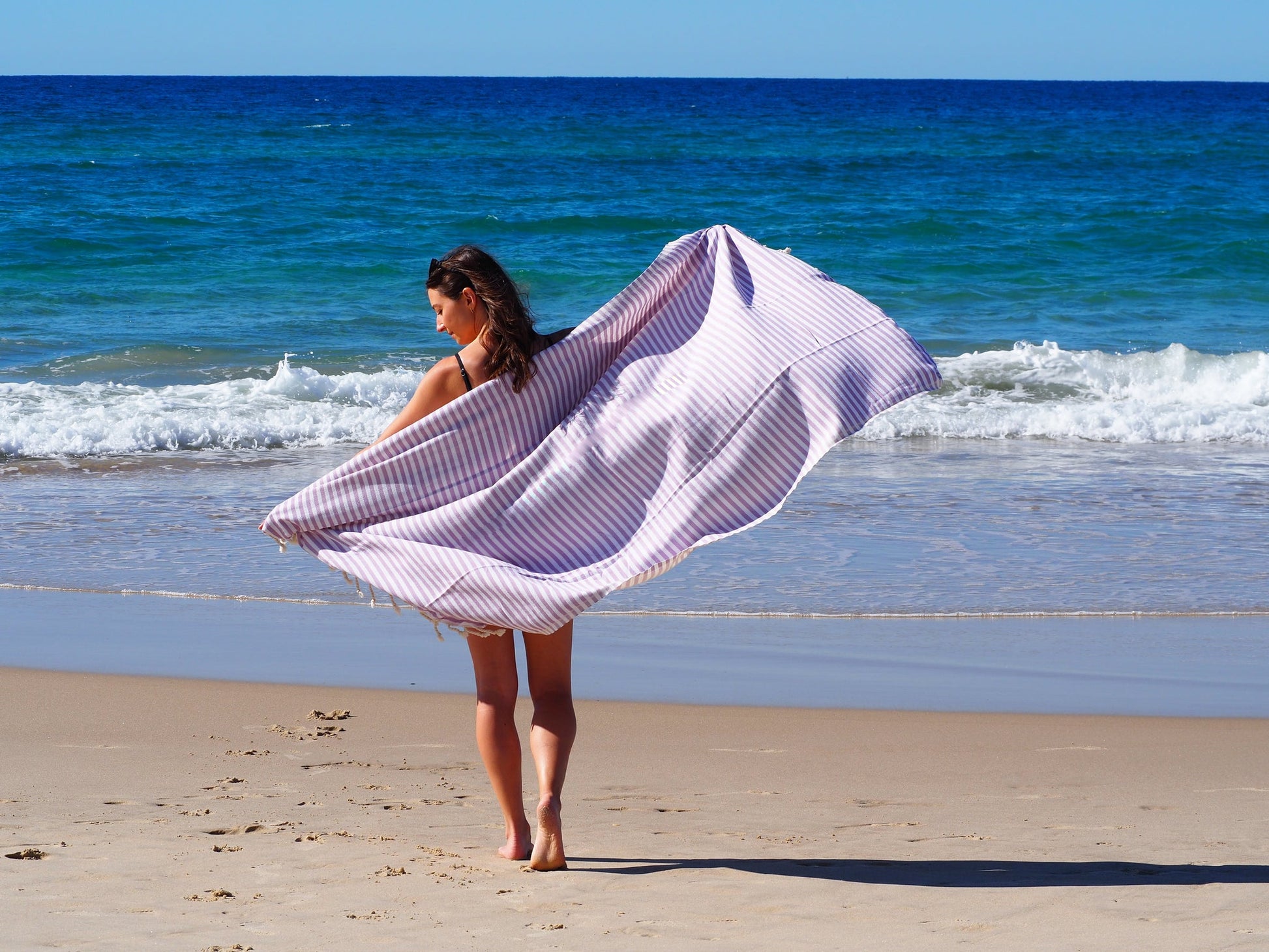 Woman enjoying the beach with a lilac Portsea Turkish cotton beach towel flowing in the breeze near the ocean.