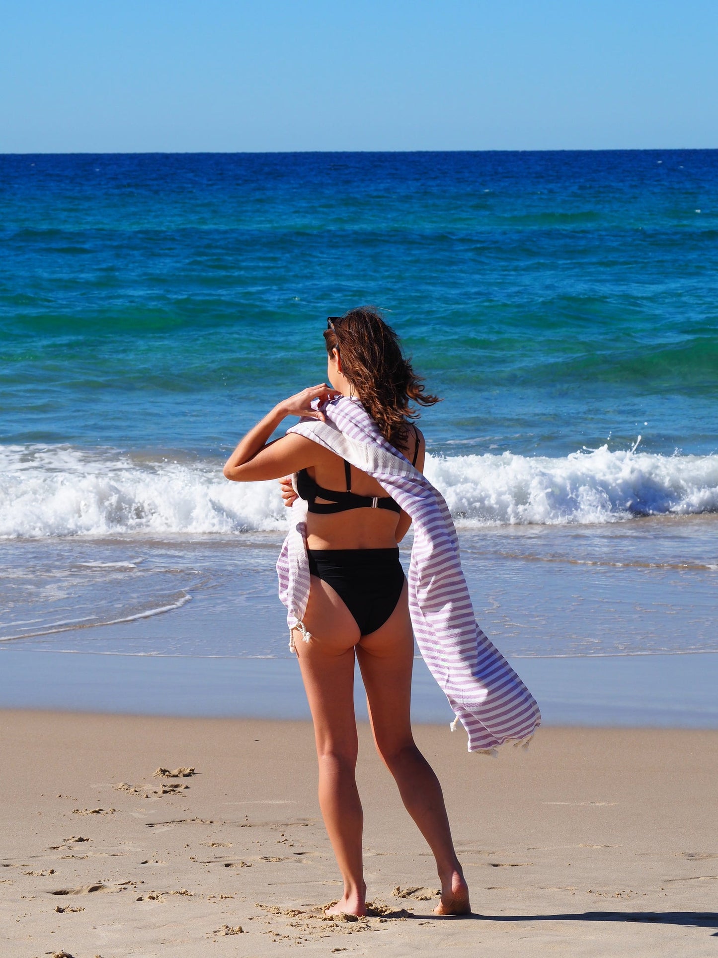 Woman wearing a black bikini holding a lilac striped beach towel on a sunny beach with ocean waves in the background.