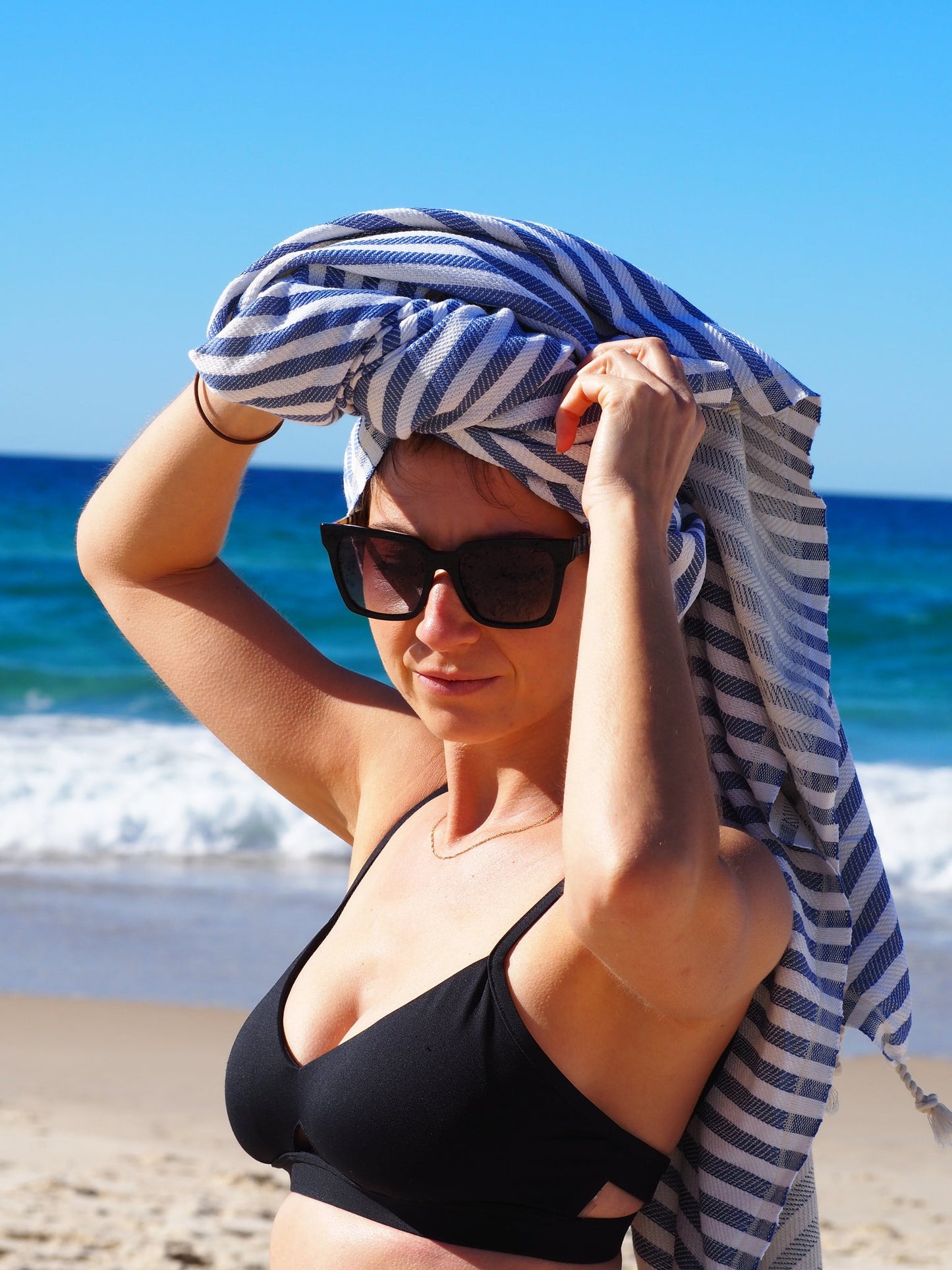 Woman in a black bikini on the beach wrapping a striped towel on her head with the ocean in the background.