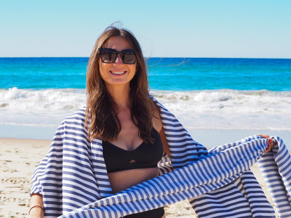 Woman in sunglasses wearing a striped beach towel, smiling on the beach with ocean waves in the background.