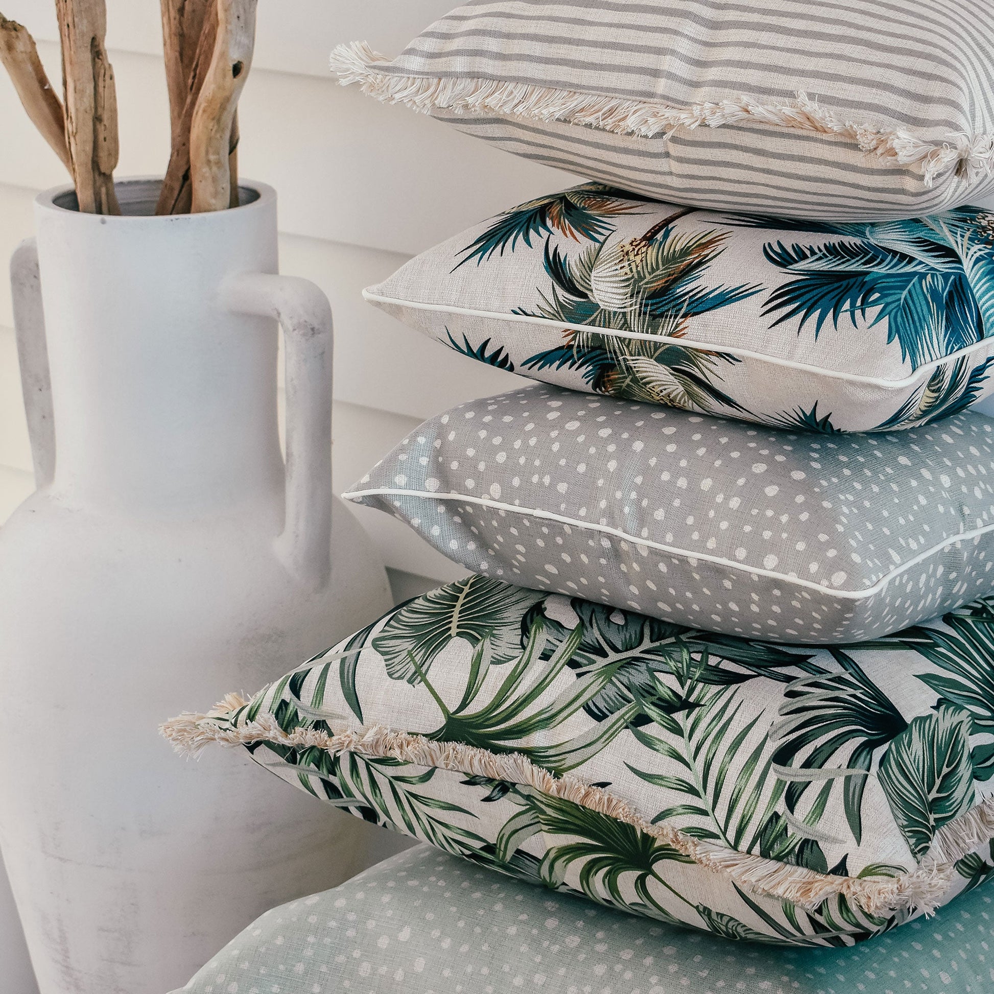 Assorted decorative cushion covers featuring tropical palm prints and textured fabrics, styled with a white vase.