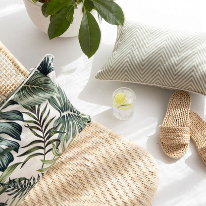 Two decorative cushion covers on a natural woven chair, with tropical and herringbone patterns, enhancing a cozy, stylish space.