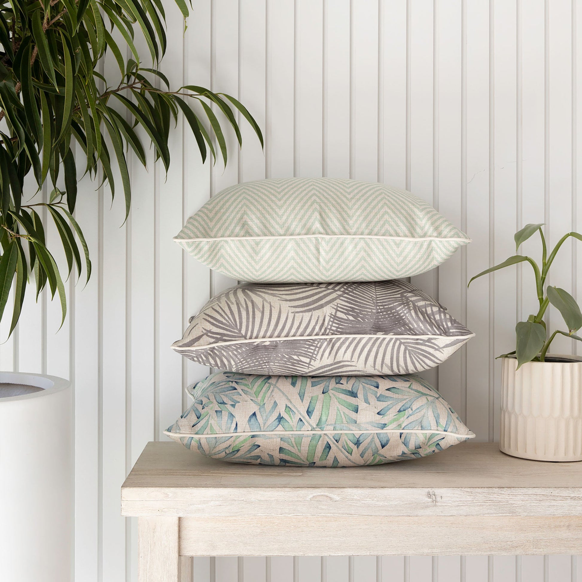 Stack of decorative cushion covers in pale mint and tropical patterns on a wooden table with plants.