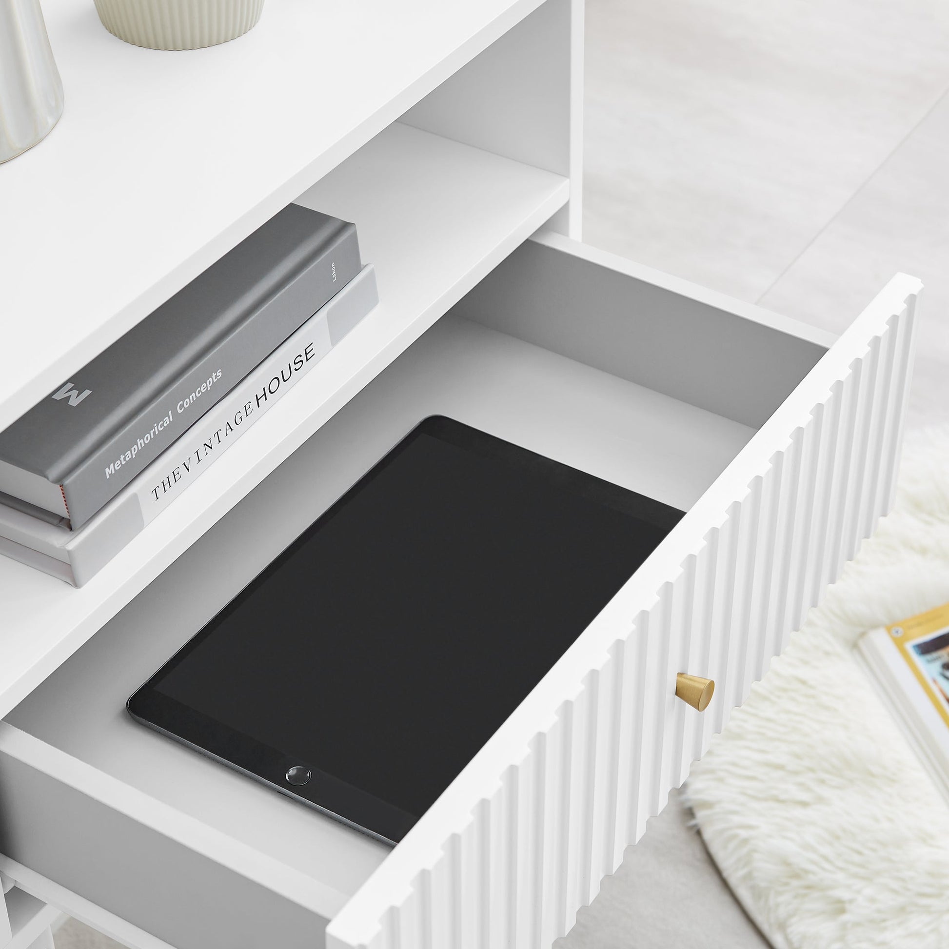 White Lucia bedside table with open drawer showing books and a tablet, featuring fluted design and satin finish.