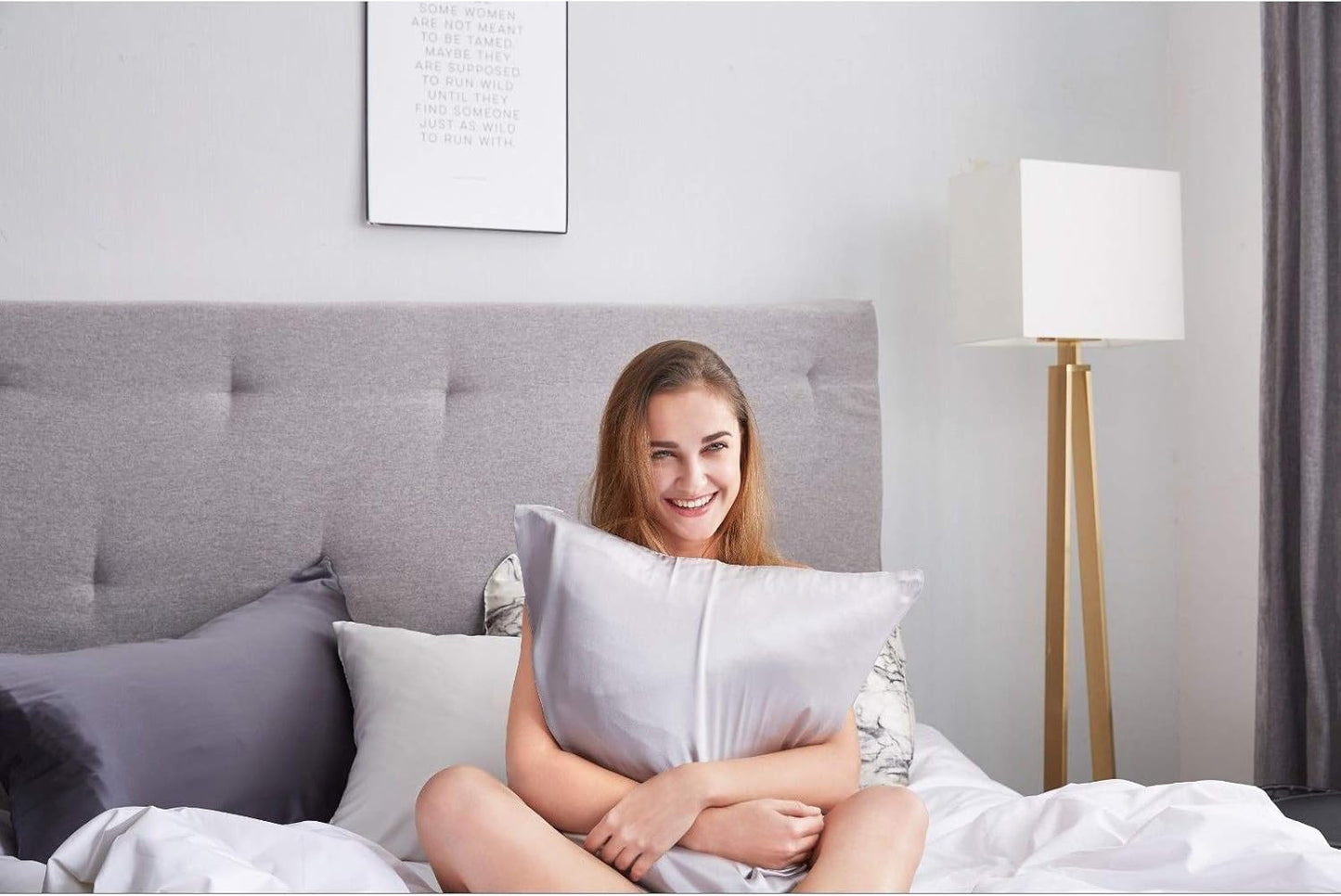 Young woman smiling while holding a luxurious silver mulberry silk pillowcase on a cozy bed.