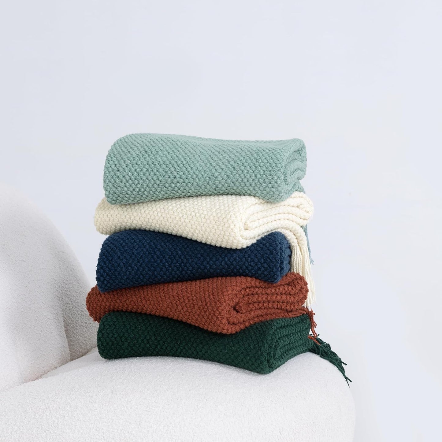 Stack of colorful knitted microfiber throw blankets, featuring green, cream, navy, rust, and forest colors on a white sofa.