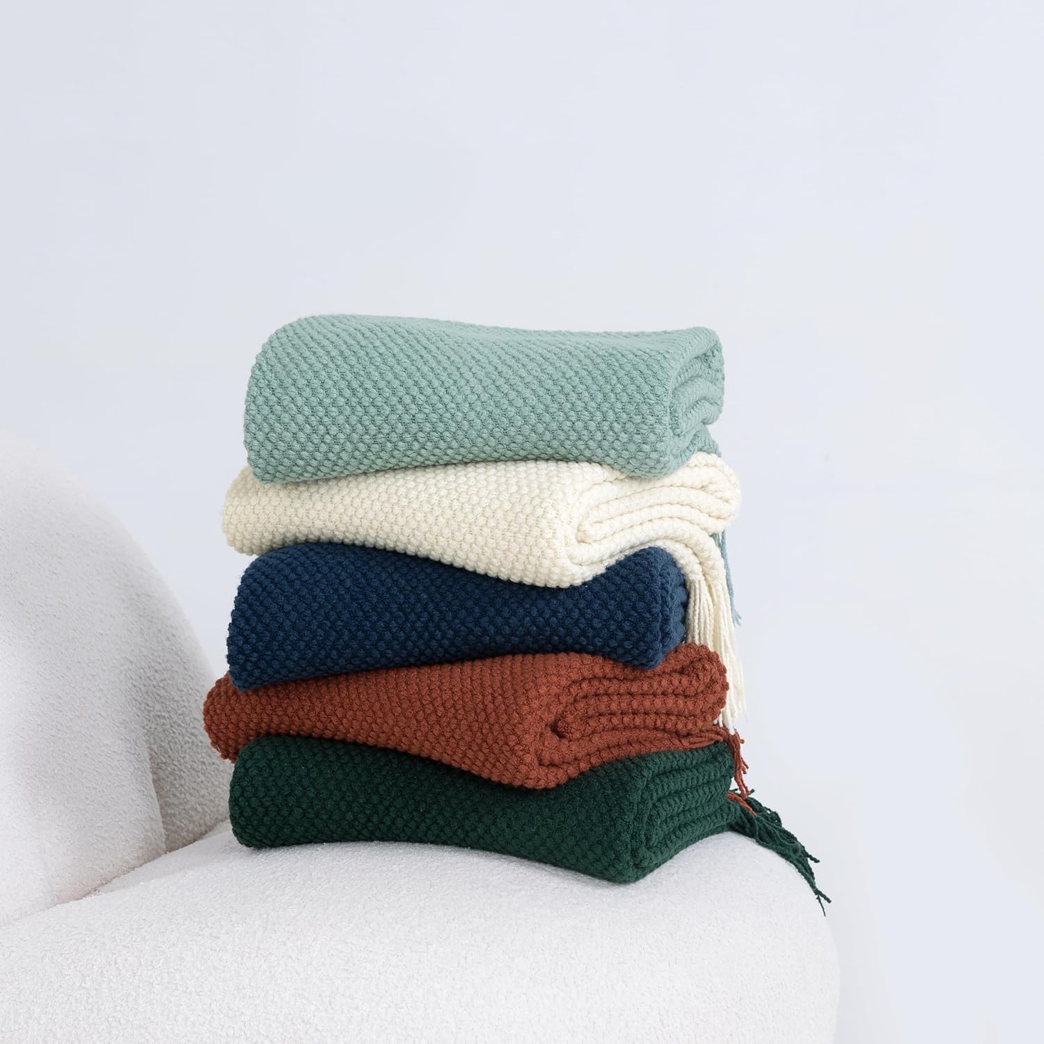 Stack of colorful knitted throw blankets in green, cream, navy, rust, and dark green on a white chair, for cozy home decor.