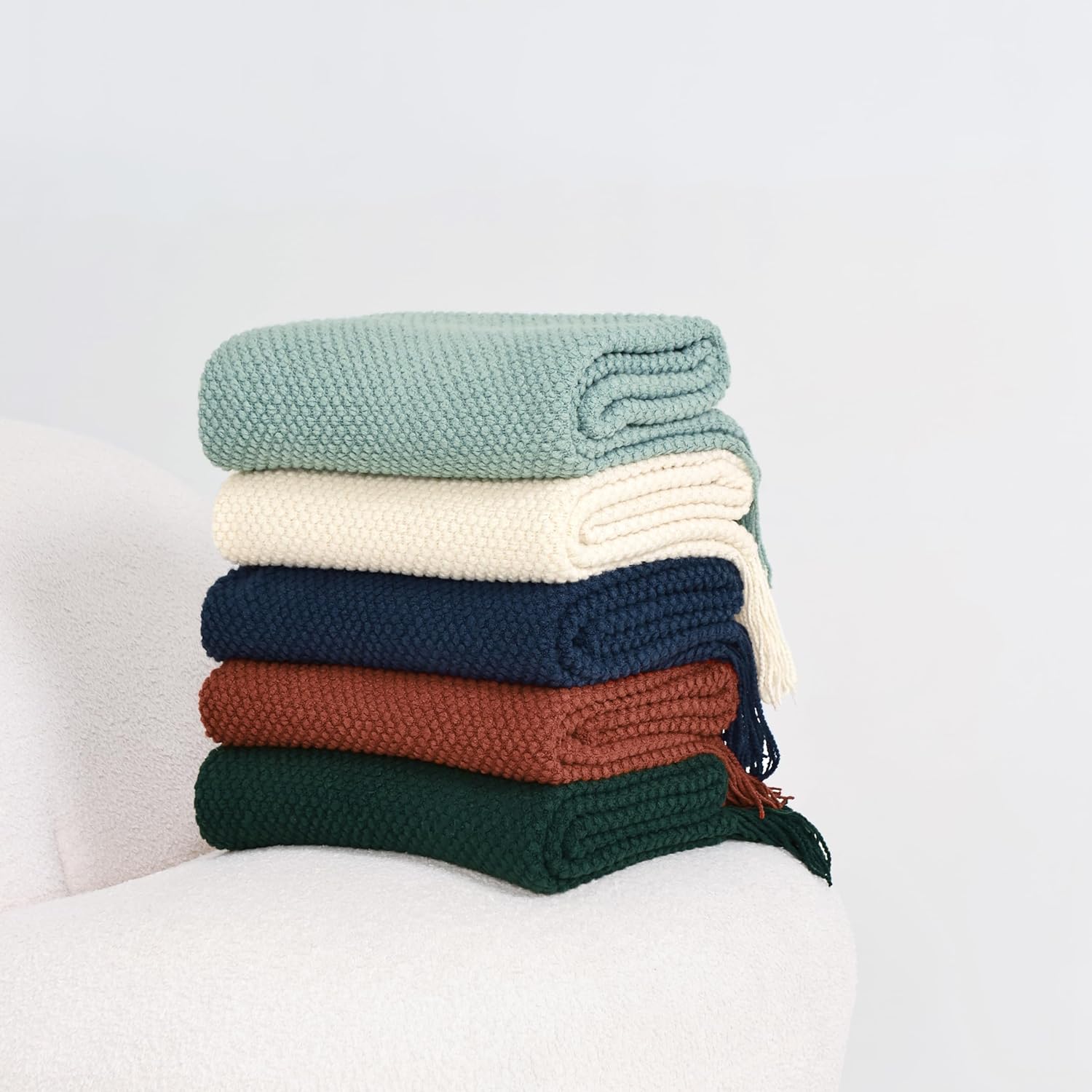 Stack of colorful Micro Plush Knitted Woven Throw Blankets including green, cream, navy, brown, and dark green on a chair.