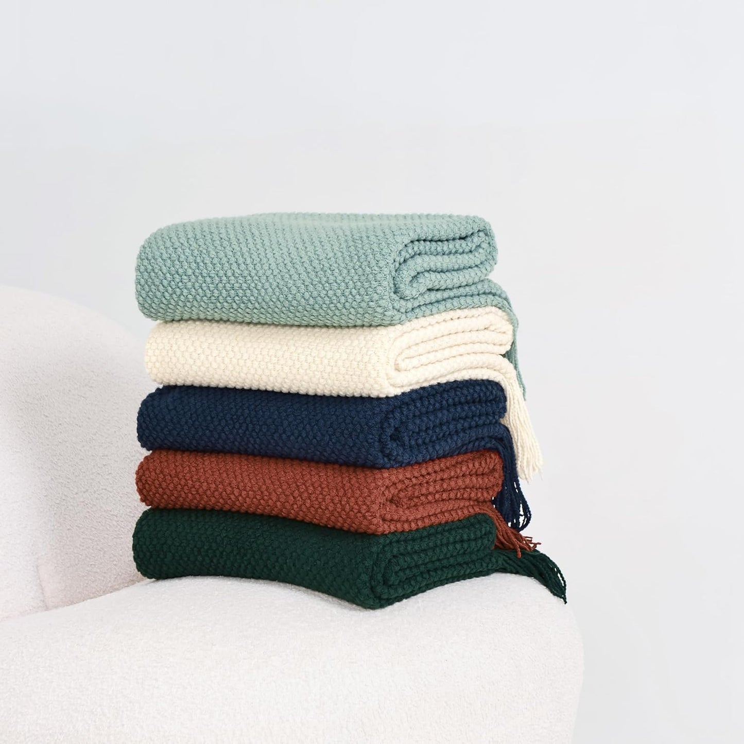 Stack of colorful knitted woven throw blankets featuring terracotta, navy, green, ivory, and sage for cozy home décor.